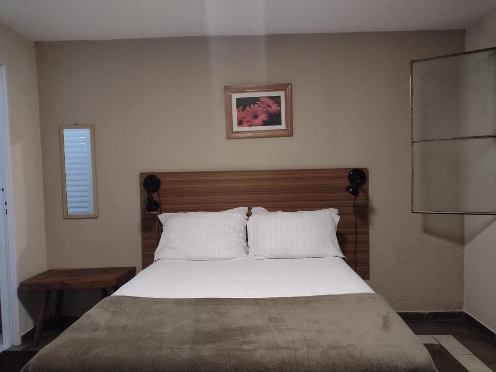 A bedroom with a wooden headboard, a bed with white linens, a small bedside table, a framed picture on the wall, and a mirror on the right.