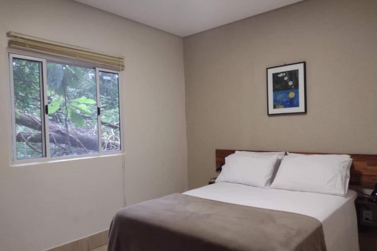 Un dormitorio con una cama, una ventana con vistas a vegetación y un cuadro en la pared.