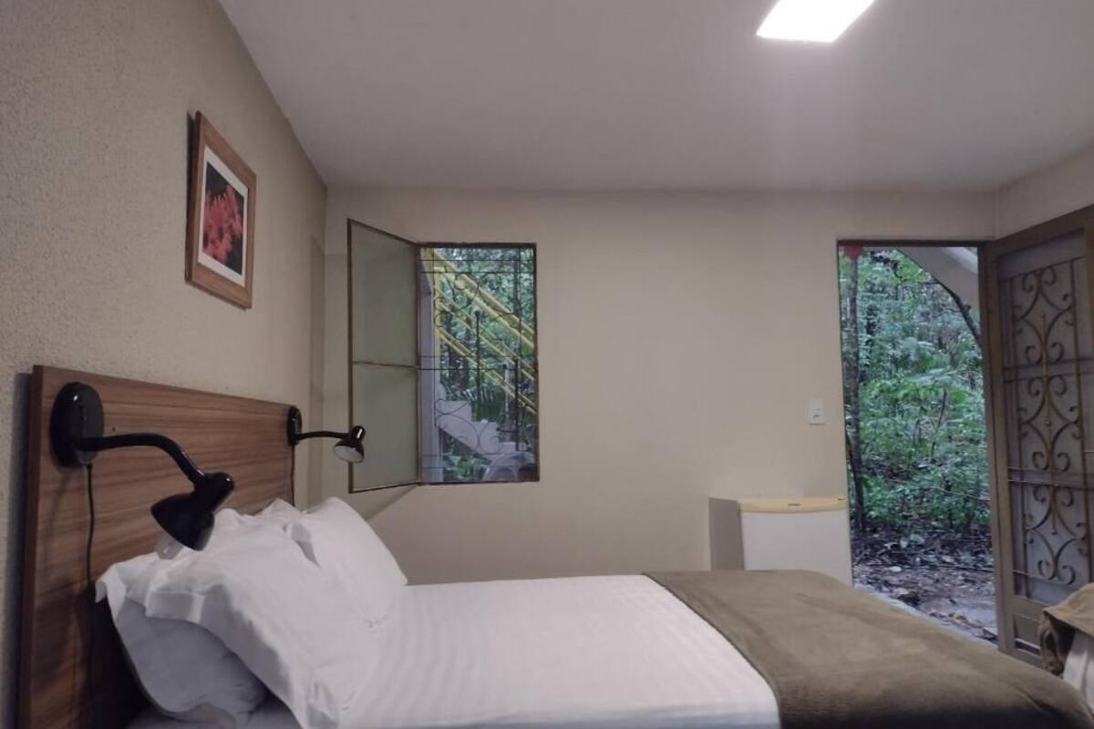 Un dormitorio con cama, mesita de noche, lámpara de pared, ventana y puerta que da a un jardín.