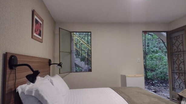 A bedroom with a bed, bedside table, nightstand, wall-mounted lamp, window, and a door leading to a garden. - Pousada Éden (Colinas do Sul)