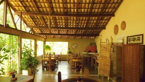 A spacious indoor area with wooden tables and chairs, a high ceiling with a bamboo roof, and large windows allowing natural light. - Pousada Éden (Colinas do Sul)