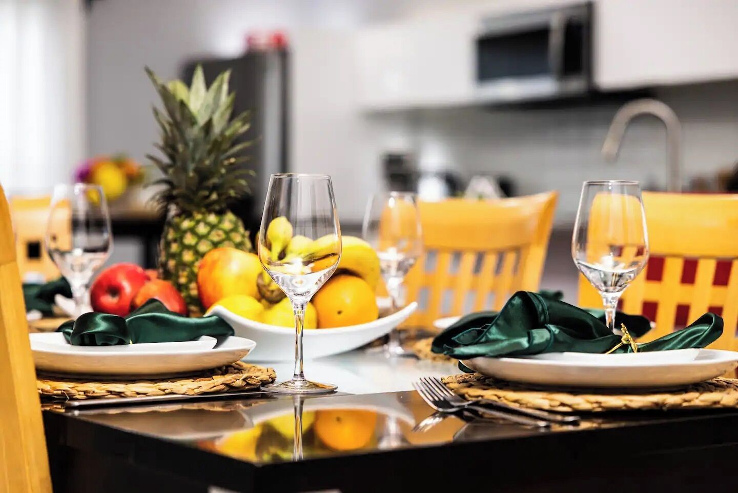 Een eettafel gedekt met borden, bestek en glazen, met een fruitschaal en een ananas op de achtergrond.