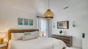 A modern hotel bedroom with a wooden headboard, a large bed with white linens, a hanging rattan light fixture, a bedside table with a lamp, a dresser, and a small plant. -     Family Pool Home-Near Disney, Food Festivals & Events- Book Now (Davenport)