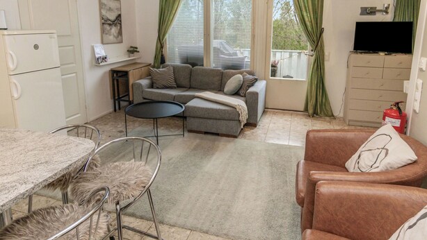 Un espace de vie compact comprenant un coin cuisine, un canapé gris, un fauteuil en cuir marron, une table à manger avec une chaise, et un téléviseur fixé au mur.