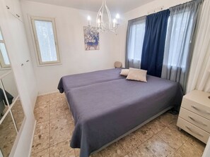 Une chambre à coucher avec un lit, une commode, un lustre, une fenêtre avec des stores et un tableau au mur.