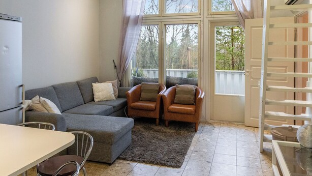 A modern living room with a grey sectional sofa, two leather armchairs, a small dining table, and a kitchen area with a refrigerator.