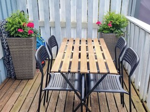 Ein Holztisch mit vier Stühlen auf einer Holzterrasse, umgeben von einem weißen Zaun und Kübelpflanzen mit rosa Blüten.
