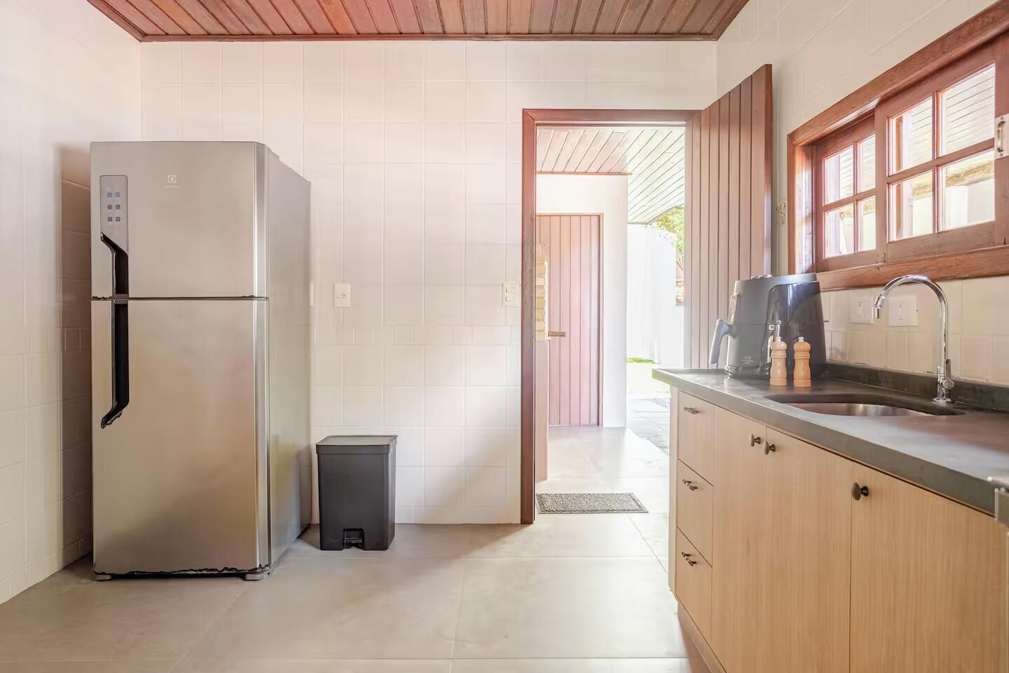 Una cocina moderna con un refrigerador de acero inoxidable, un pequeño bote de basura, armarios de madera y una ventana con persianas.