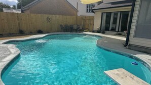 Una piscina con borde curvo, rodeada por una valla de madera y un patio con sillas.