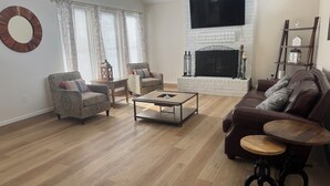 Una sala de estar con chimenea, un sofá, dos sillones, una mesa de centro y un televisor montado en la pared.