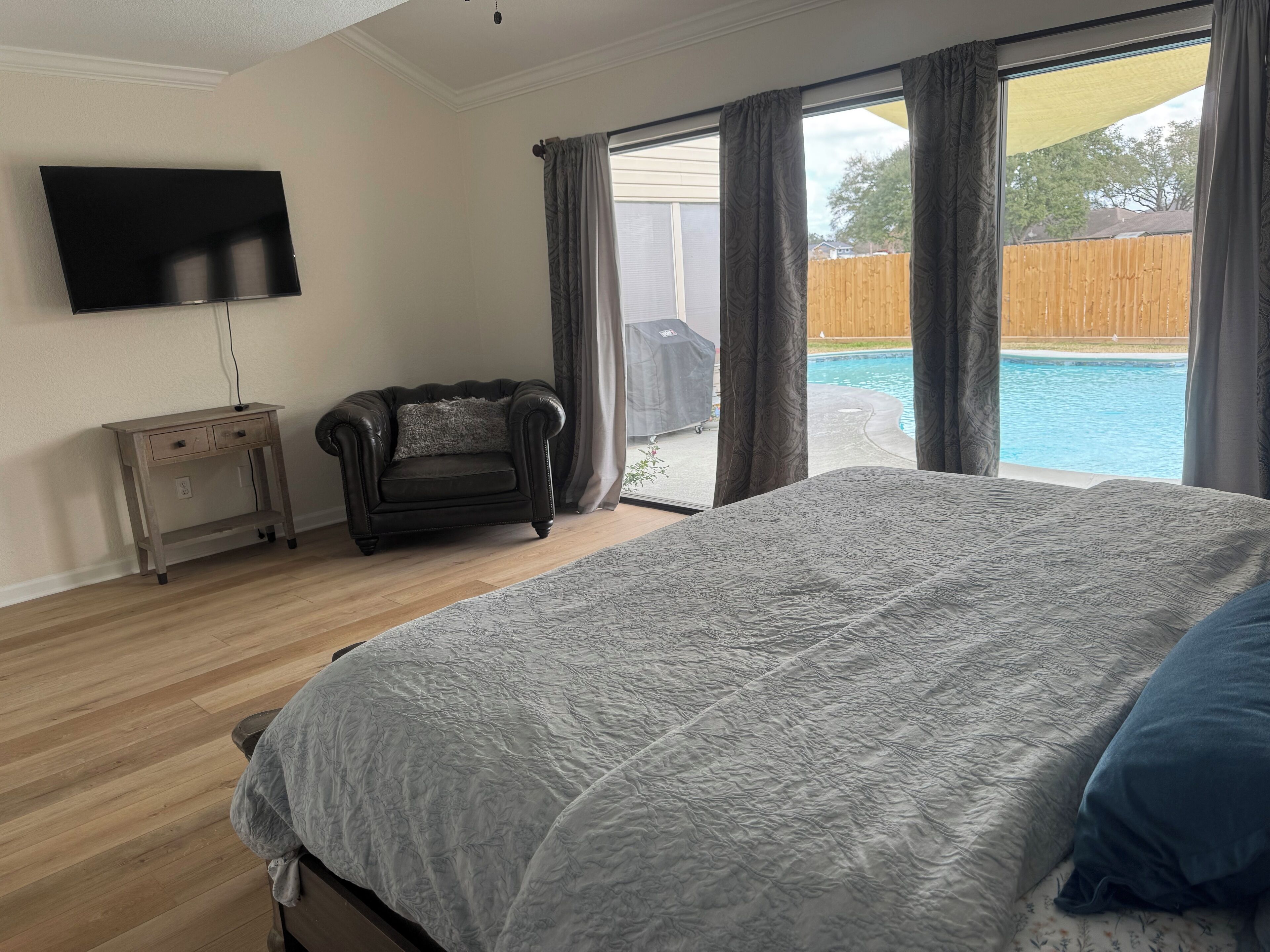 A bedroom with a large bed, a TV on the wall, a small sofa, and a view of a backyard with a pool.