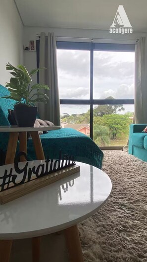 A room with a bed, a potted plant, a round table, and a view of the outdoors through large windows. - Conforto em Gramado  GRMEVE28 (Gramado)
