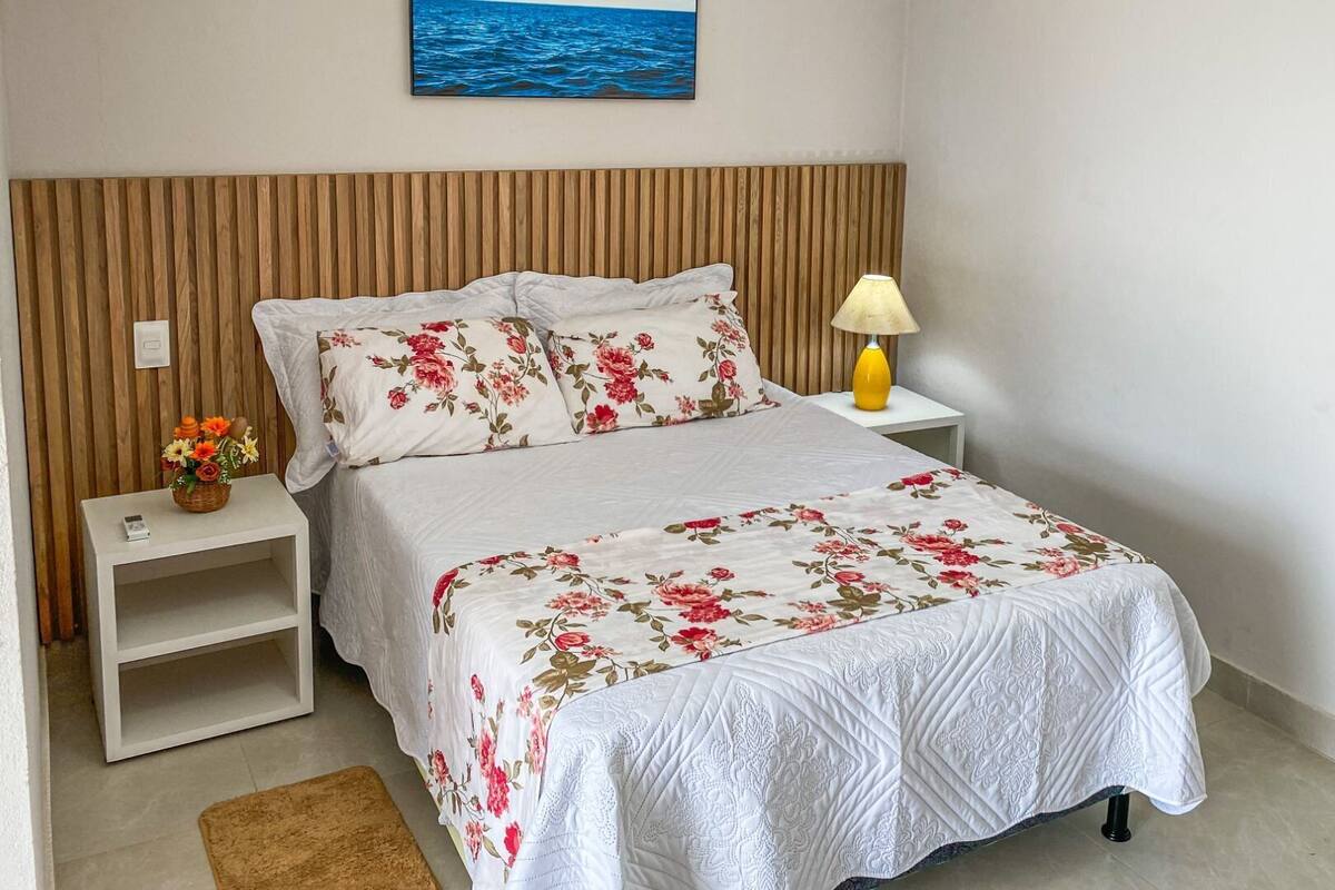A neatly arranged bedroom with a wooden headboard, floral bedding, a bedside lamp, and a small table with a vase of flowers.