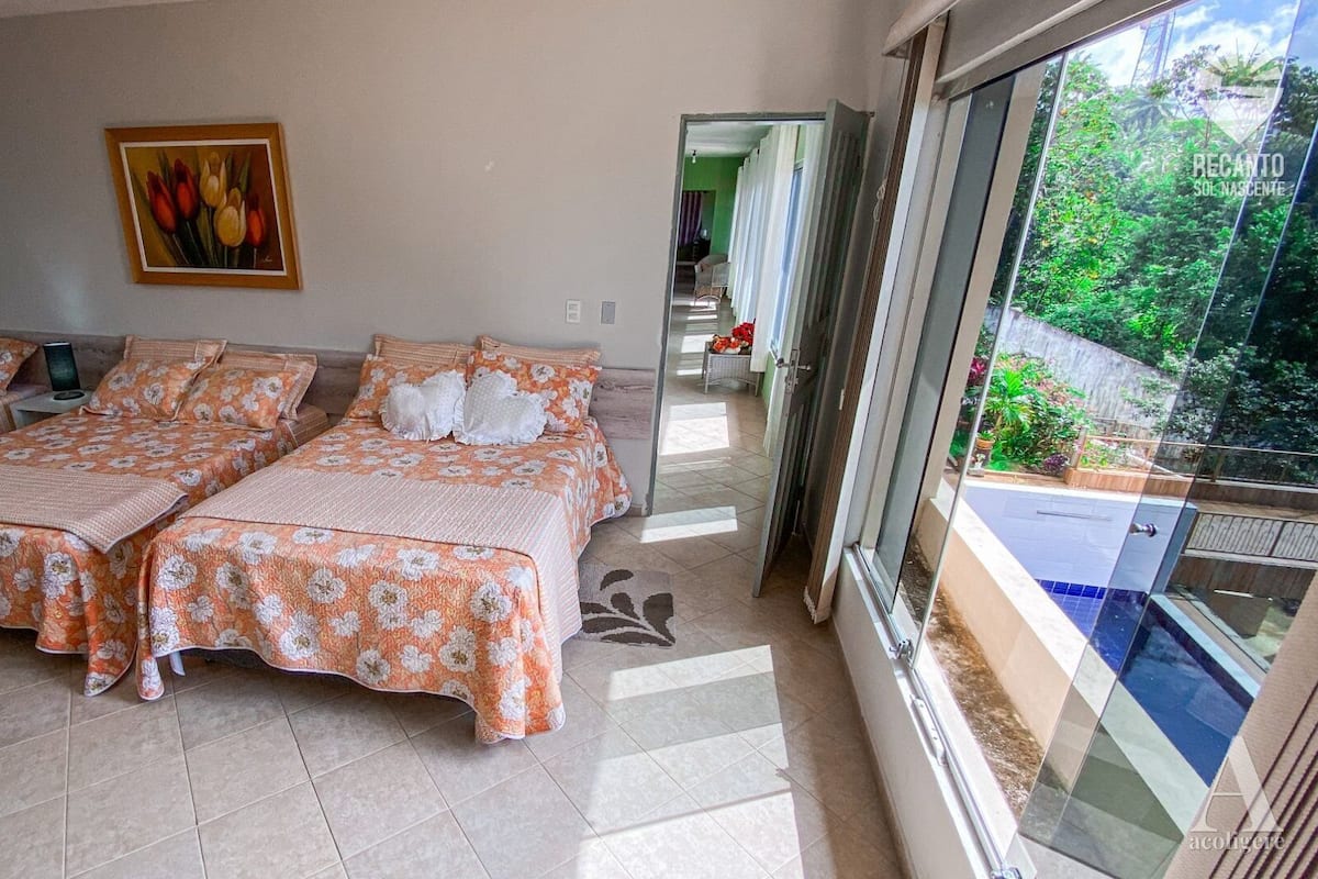 A hotel room with two beds, a painting on the wall, and a balcony with a view of greenery.