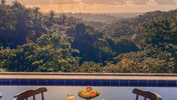 A table with breakfast items and chairs on a terrace with a view of a mountainous landscape and a pool. - Pousada Recanto Sol Nascente (Rio Formoso)
