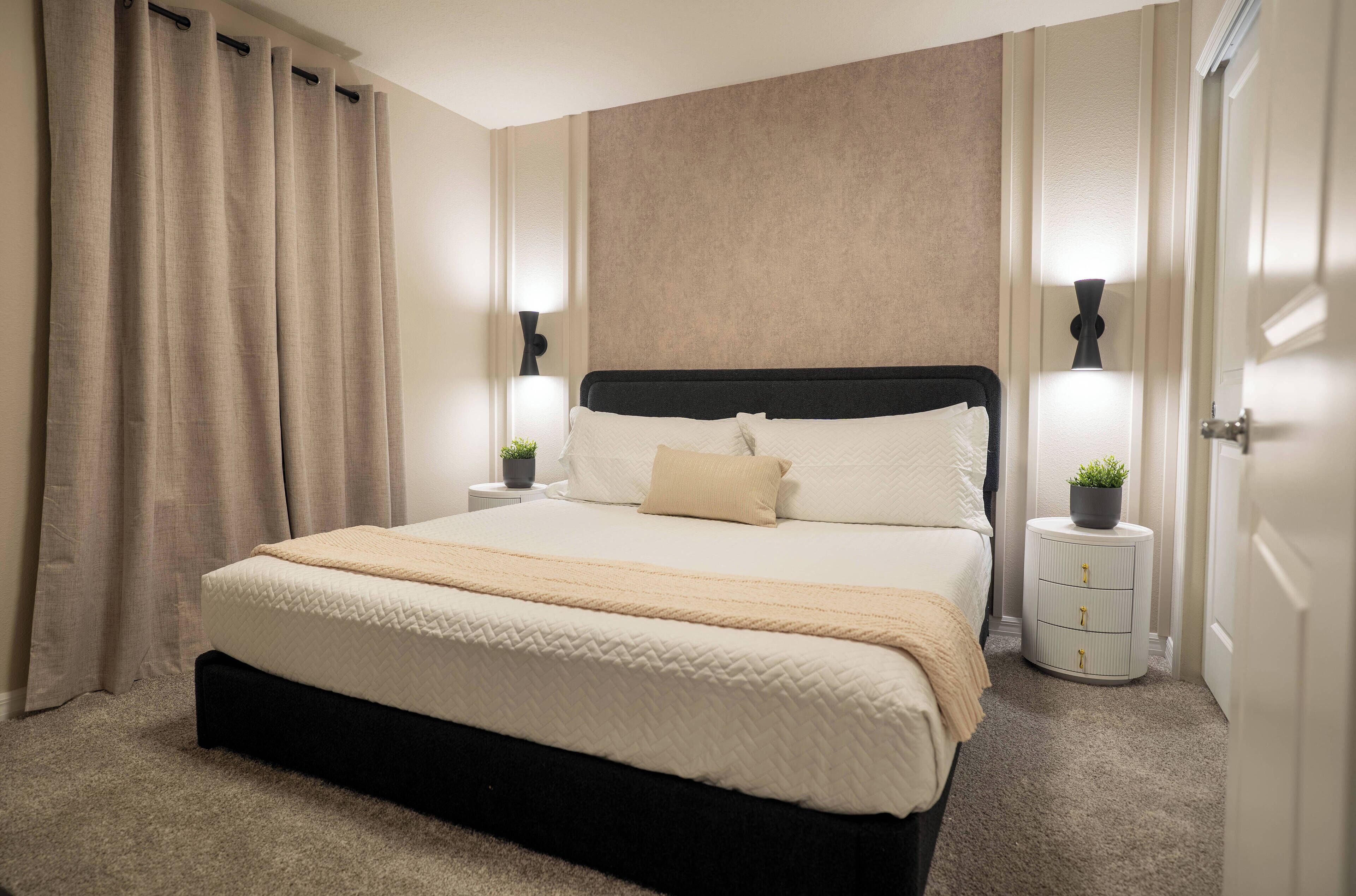 Cama bem arrumada com cabeceira, duas mesas de cabeceira com abajures e uma pequena planta em uma das mesas.
