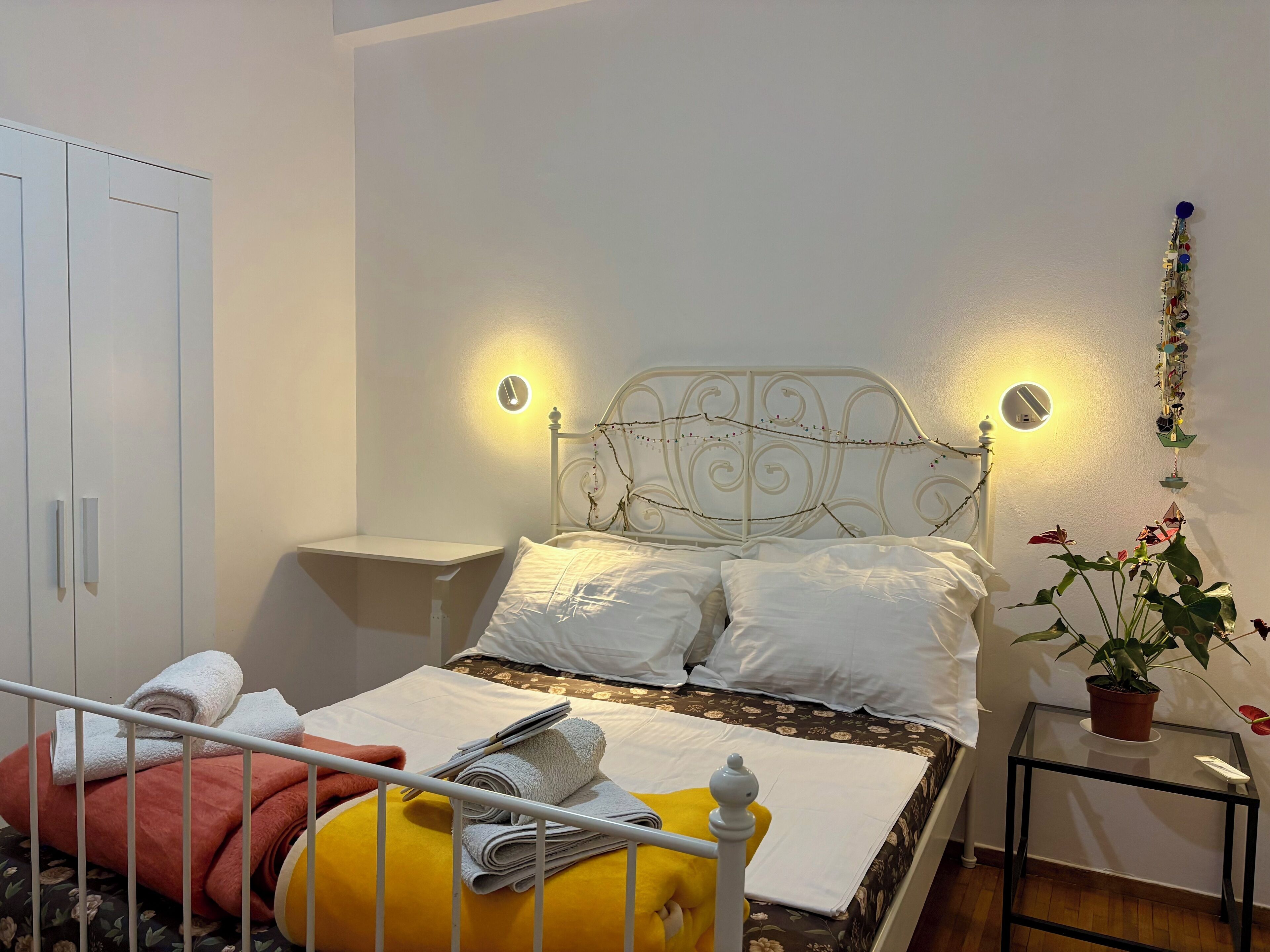 A neatly made bed with white linens, a bedside table with a lamp, a wardrobe, and a potted plant.