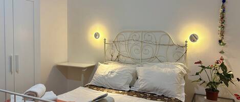 A neatly made bed with white linens, a bedside table with a lamp, a wardrobe, and a potted plant.