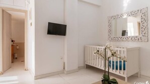 Una habitación con paredes blancas, un televisor montado en la pared, una cuna blanca con un espejo decorativo encima y una planta en maceta en primer plano.