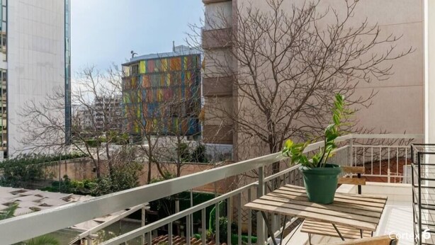 Un balcón con una mesa de madera y una planta en maceta, con vistas a un paisaje urbano con edificios modernos y un edificio colorido al fondo.