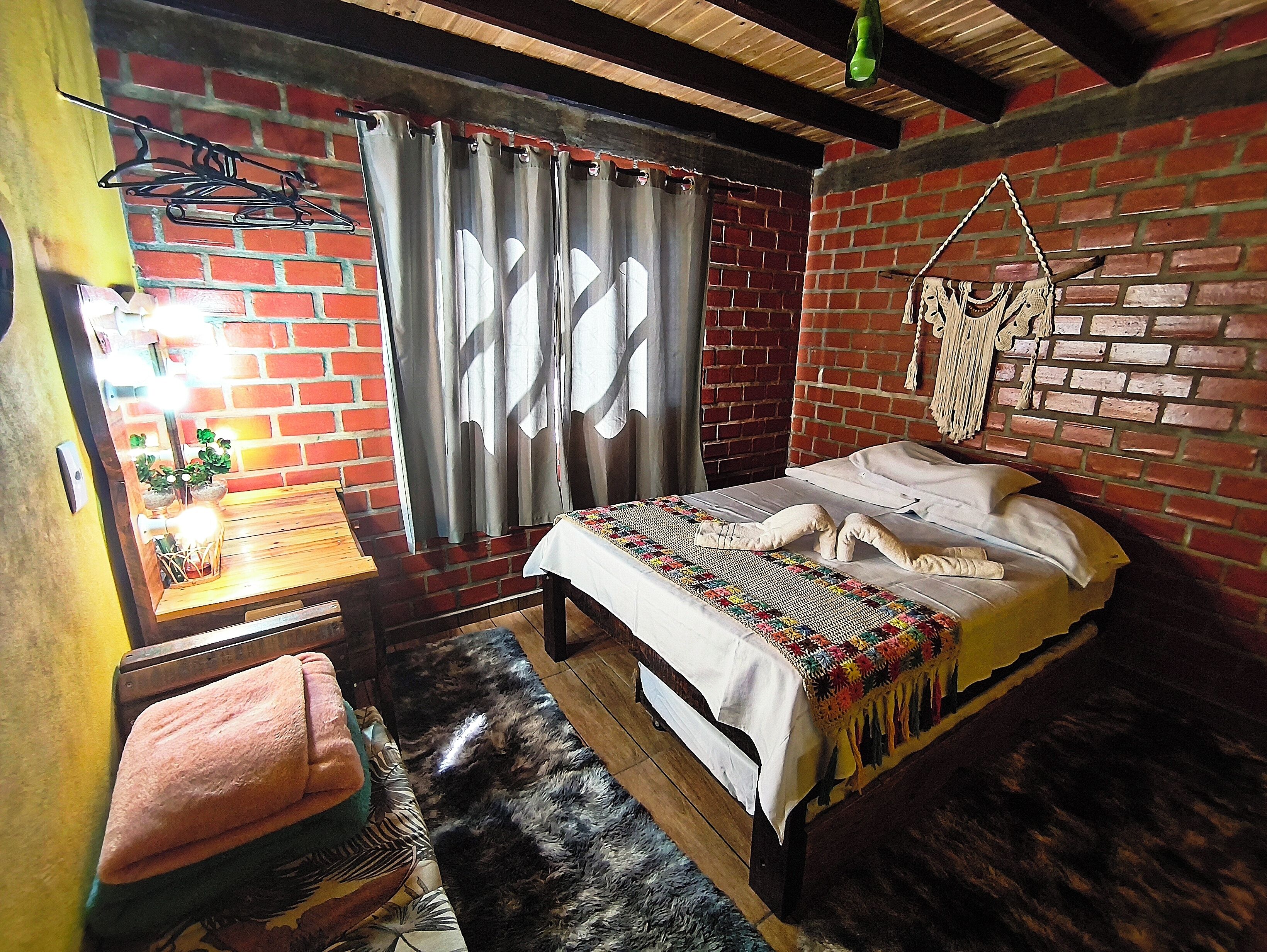 A room with a brick wall, a bed with a colorful blanket, a wooden bench, and a window with curtains.