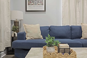 Un salon avec un canapé bleu, une lampe blanche, un tableau encadré au mur et une table basse sur laquelle se trouvent une plante et un panier.