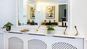 Une console blanche, surmontée d’un grand miroir qui reflète un coin repas. Sur la console, on trouve deux plantes en pot, un petit vase blanc et deux bougeoirs.