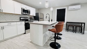 Una cocina moderna con armarios blancos, una isla central, microondas y fregadero. Hay dos taburetes de bar y una mesa de comedor con sillas.