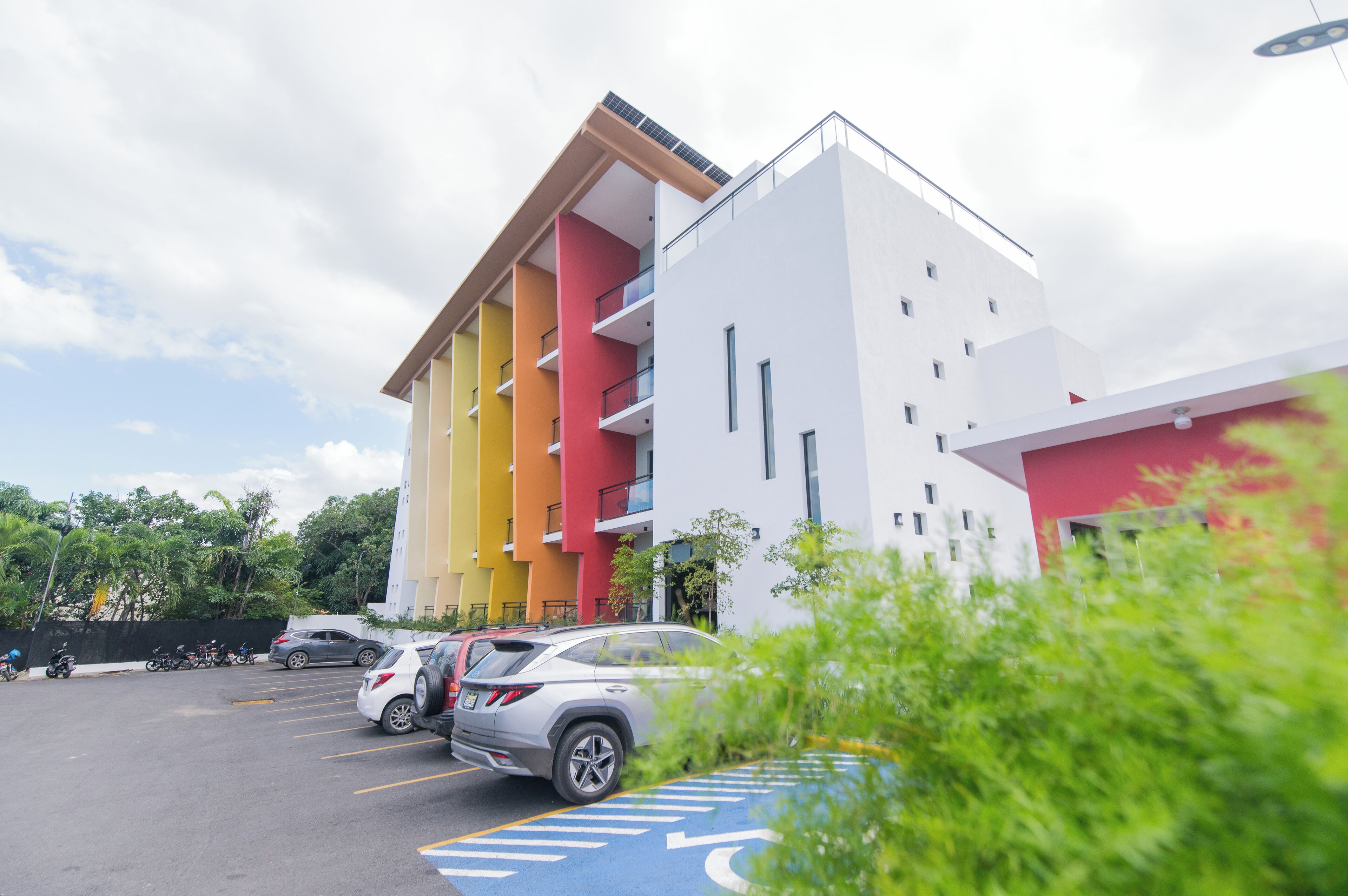 Um edifício de vários andares com design moderno, apresentando uma combinação de cores branco, amarelo e vermelho. Vários carros estão estacionados em frente ao edifício, e uma vaga de estacionamento marcada com o símbolo de cadeira de rodas.