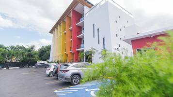 Um edifício de vários andares com design moderno, apresentando uma combinação de cores branco, amarelo e vermelho. Vários carros estão estacionados em frente ao edifício, e uma vaga de estacionamento marcada com o símbolo de cadeira de rodas.