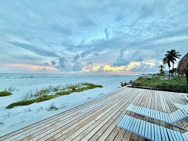 Eine Holzterrasse mit Liegestühlen mit Blick auf einen Strand und das Meer, Palmen sind zu sehen.