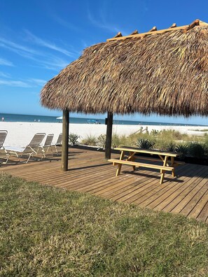 Eine Holzterrasse am Strand mit Liegestühlen und einem Picknicktisch unter einem Reetdach.