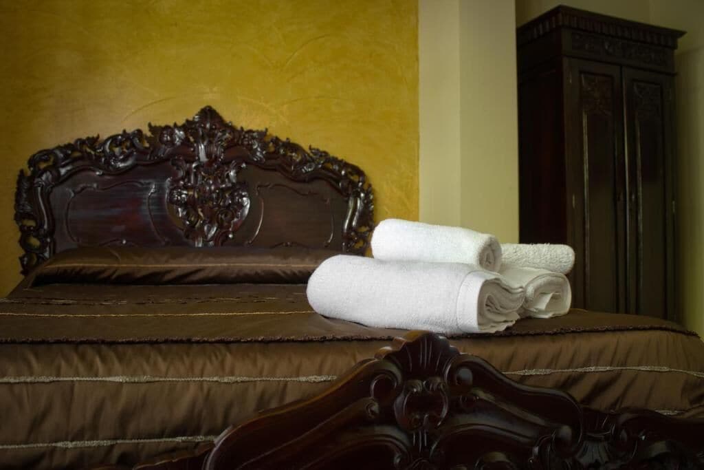 Una cama con cabecera oscura y colcha marrón, acompañada de dos toallas blancas enrolladas.