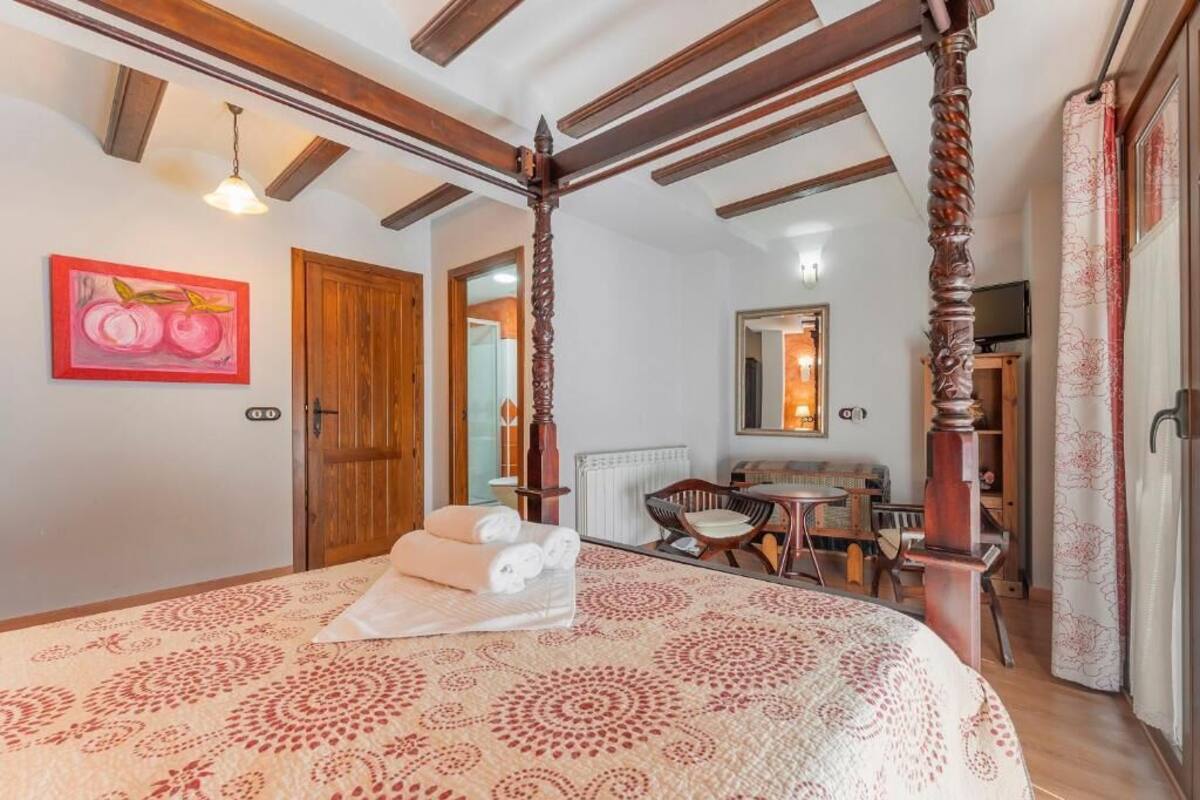 A bedroom with a four-poster bed, a wooden ceiling, a painting of fruit, a small table with chairs, and a bathroom visible through an open door.