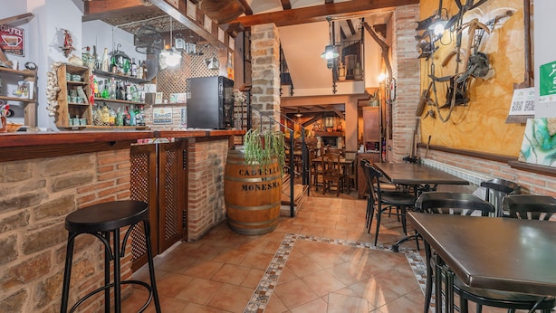 A rustic bar area with wooden beams, a stone wall, and a large barrel. There are bar stools, tables, and a variety of bottles and glasses on shelves.
