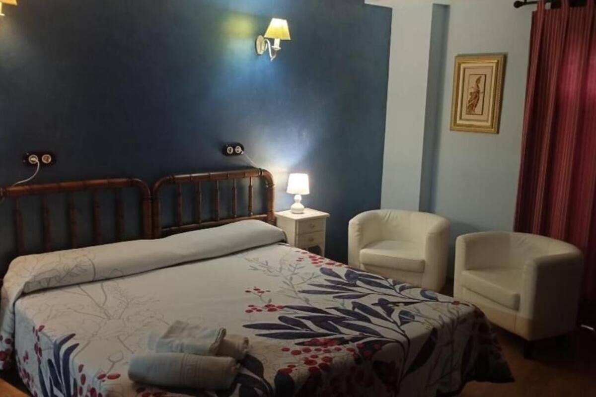 A bedroom with a bed, two bedside tables, a nightstand, two chairs, a lamp, and a framed picture on the wall.