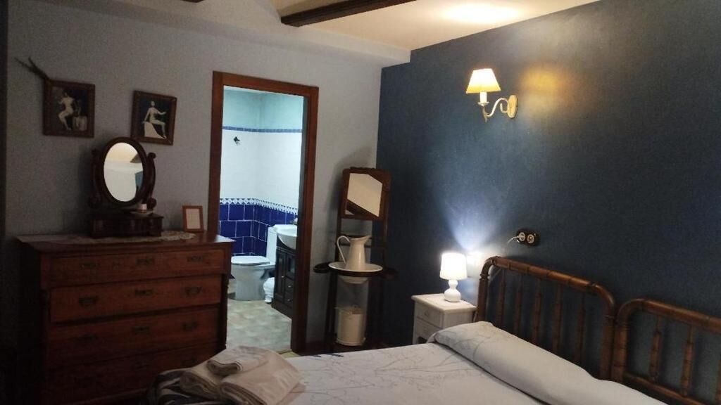 A bedroom with a bed, dresser, bedside tables, and a mirror. There is a door leading to a bathroom with a blue and white tiled wall.