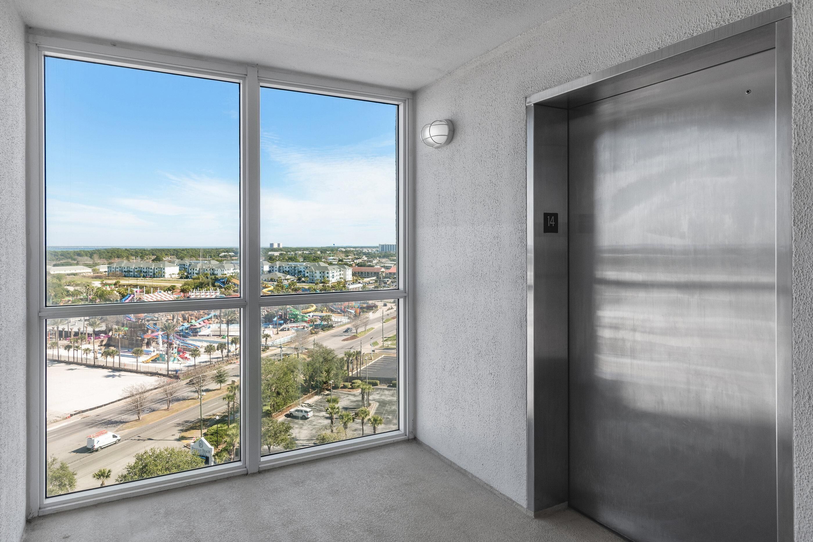 A room with a large window offering a view of a cityscape, an elevator, and a textured wall.