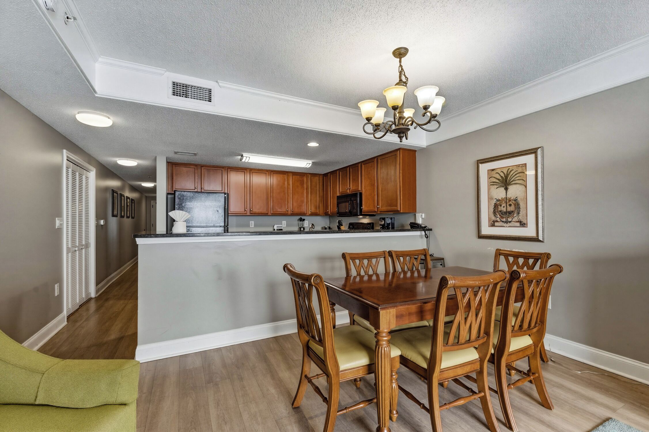 A dining area with a wooden table and chairs, a chandelier, and a kitchen in the background.
