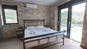 Een slaapkamer met een stenen muur, een bed met metalen frame, een nachtkastje, een raam en een balkon met uitzicht op groen.
