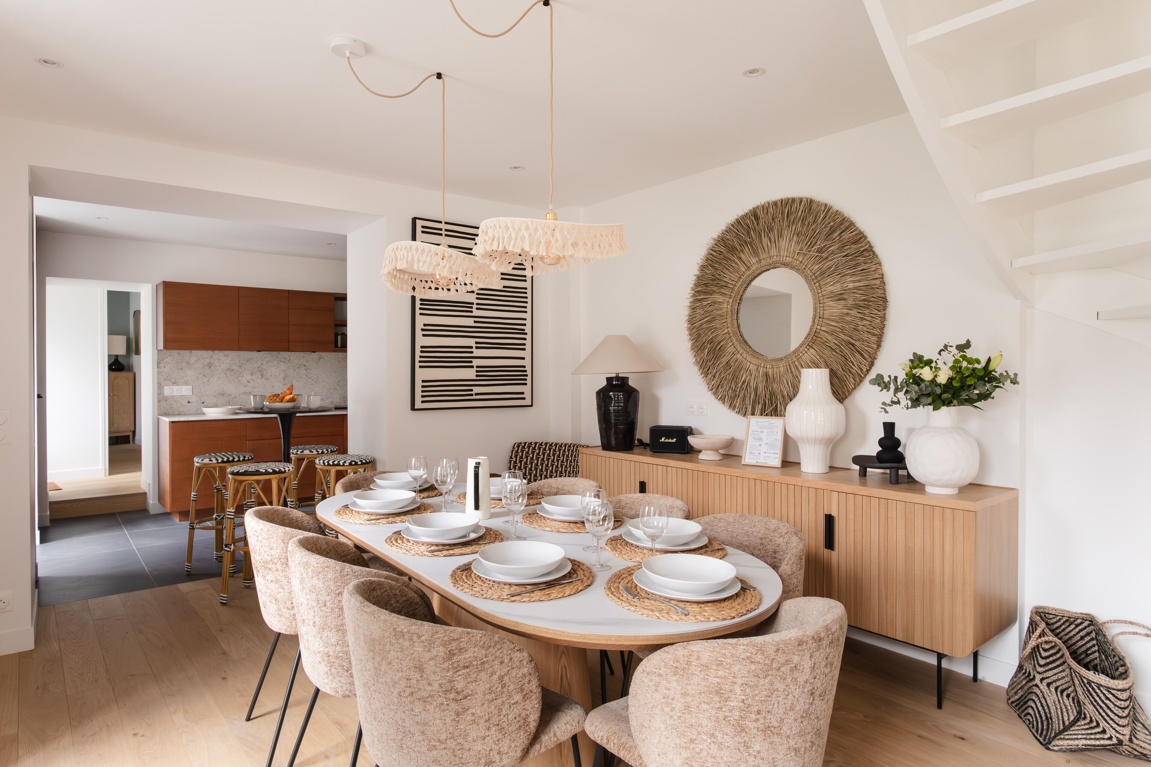 A modern dining area with a wooden table set for six, surrounded by upholstered chairs. A kitchen is visible in the background, and a large mirror hangs on the wall.