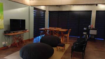 A living room with a television, a dining table, chairs, bean bags, and a small table with items on it.