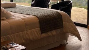 A bedroom with a large bed, two chairs, and a view of a mountainous landscape through large windows. - Pousada Refúgio dos Vinhedos (Vale dos Vinhedos)