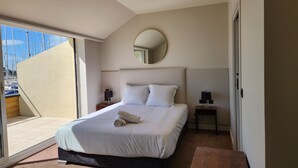 A bedroom with a bed, two bedside tables, a round mirror, and a view of boats through a large window. - Les Trois Voiles (Port-Saint-Louis-du-Rhône)