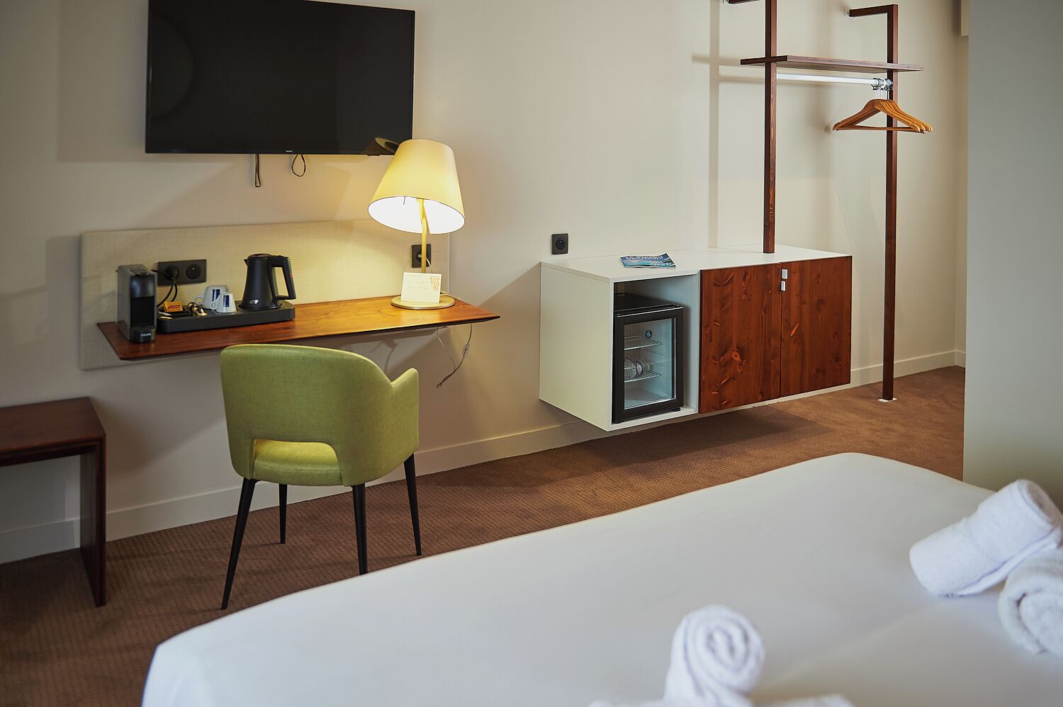 A hotel room with a bed, a desk with a lamp, a mini-fridge, a TV, and a wardrobe.