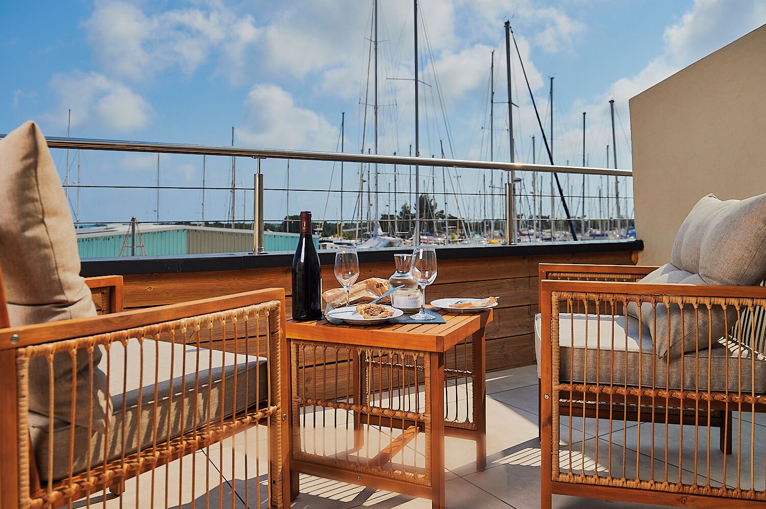 Una terraza con mobiliario de mimbre, una mesa dispuesta con platos, vasos y una botella, con vistas a un puerto deportivo lleno de barcos.