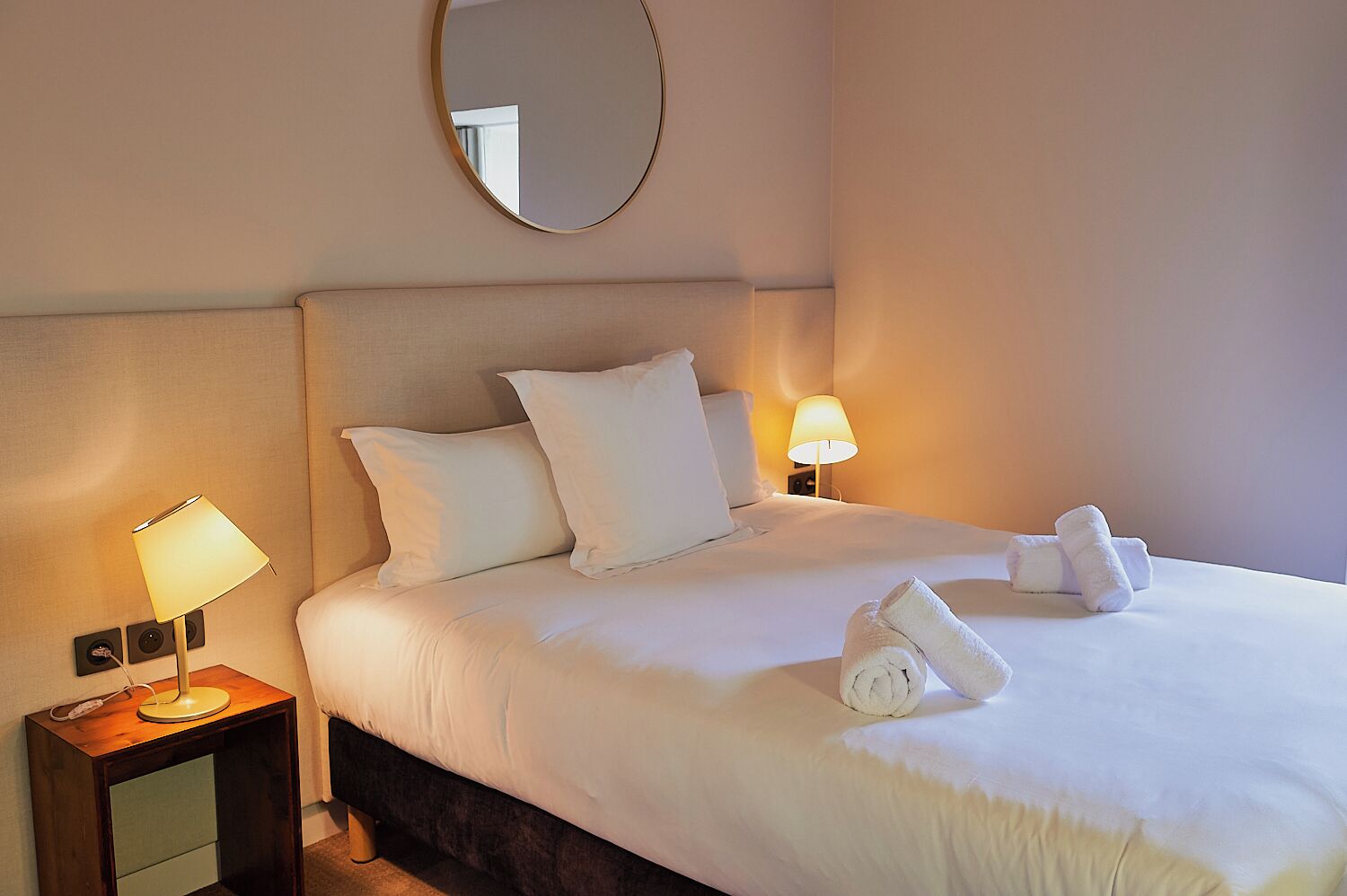 Une chambre d’hôtel avec un lit bien fait, deux lampes de chevet, un miroir rond au-dessus du lit et une table de chevet en bois avec une lampe et un petit haut-parleur.