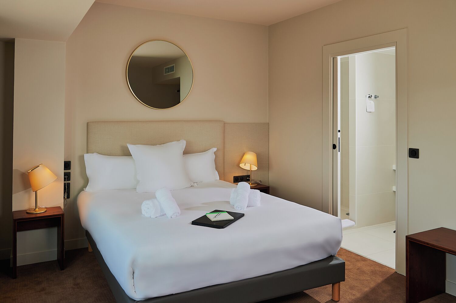 A hotel room with a large bed, two bedside lamps, a round mirror, and a door leading to a bathroom.