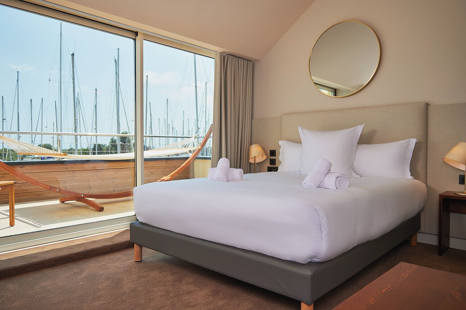 Habitación de hotel con una cama grande, una hamaca, vista a veleros y un espejo redondo en la pared.