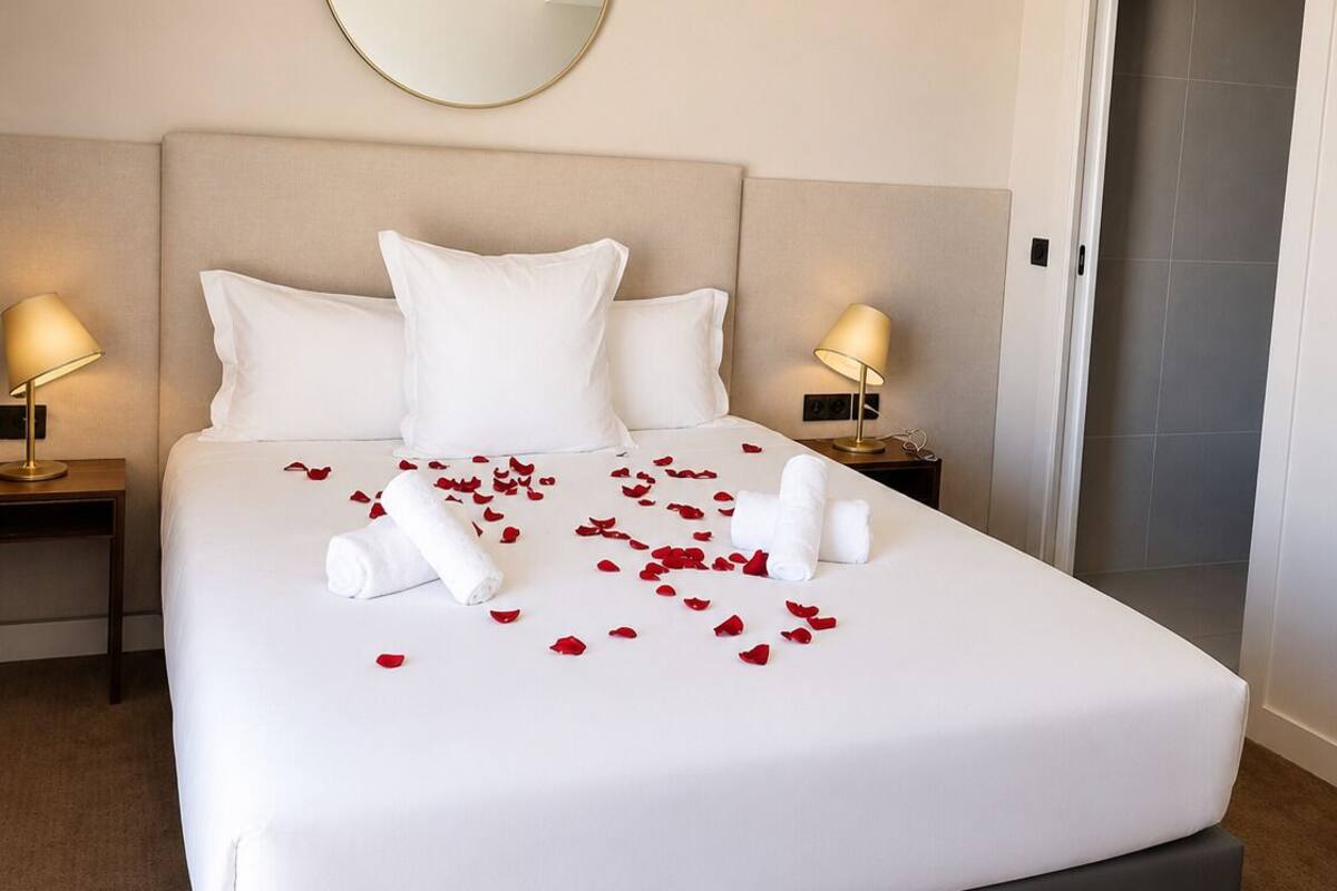 Ein ordentlich bezogenes Bett mit weißen Leinen, ein runder Spiegel darüber und rote Rosenblätter, die auf dem Bett verteilt sind.
